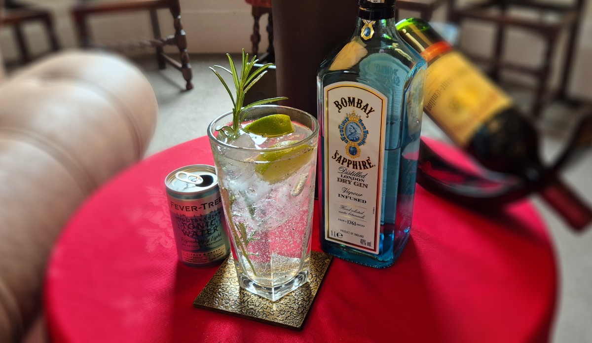 A tall glass filled with ice, gin and tonic and containing lime wedges and a long sprig of rosemary, on a table between a bottle of Bombay Sapphire gin and a can of Fever Tree Mediterranean tonic water